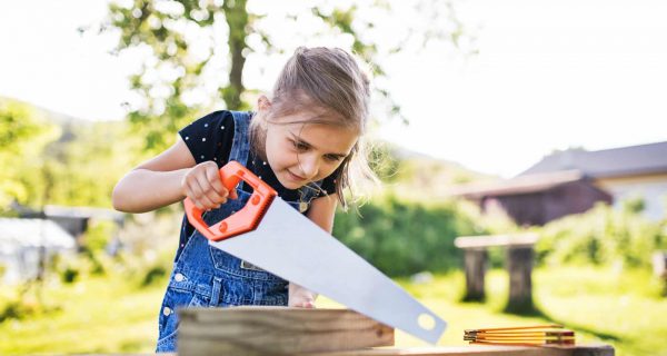 A small girl with a saw outside, making a wooden birdhouse.